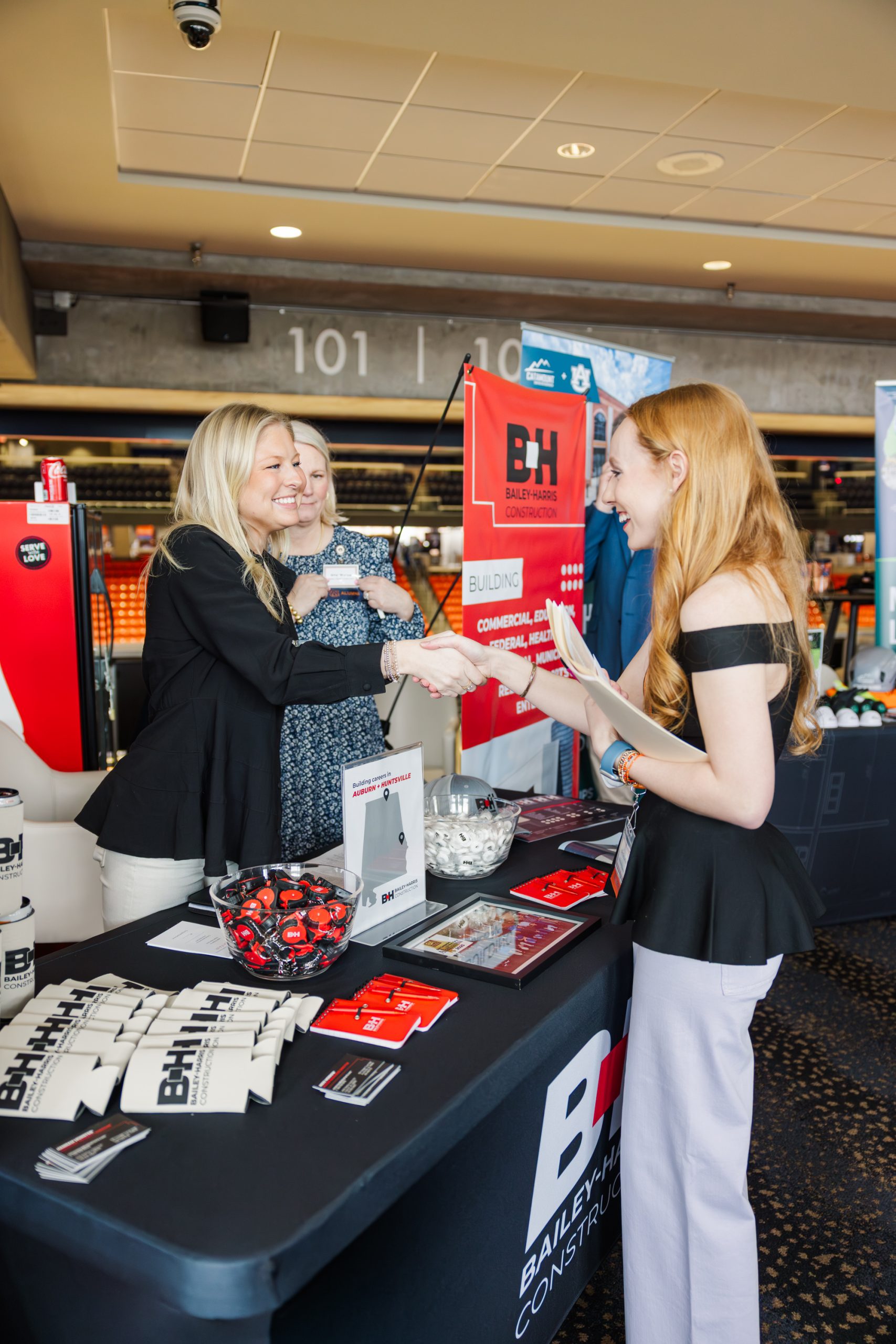 Two women stand behind a promotional table with Bailey Harris Construction branding, greeting a third woman at an indoor event.