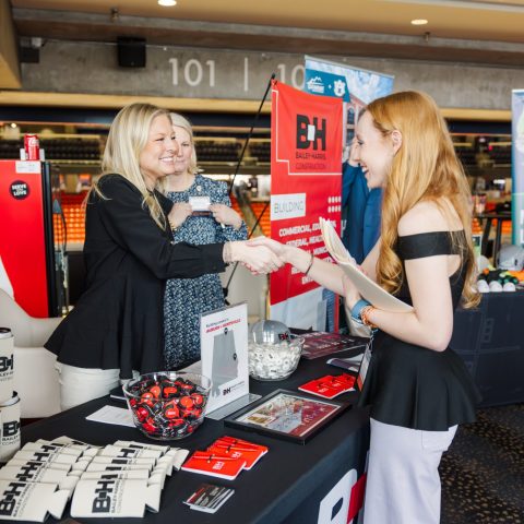 Two women stand behind a promotional table with Bailey Harris Construction branding, greeting a third woman at an indoor event.
