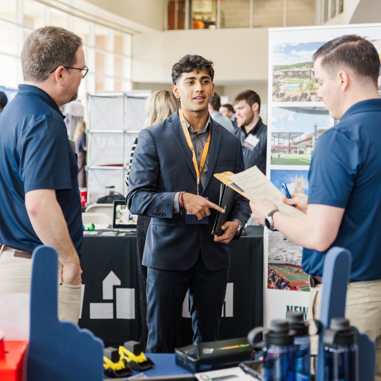 A young man in a suit speaks with two men at a business expo booth, with promotional items and informational displays on the table.