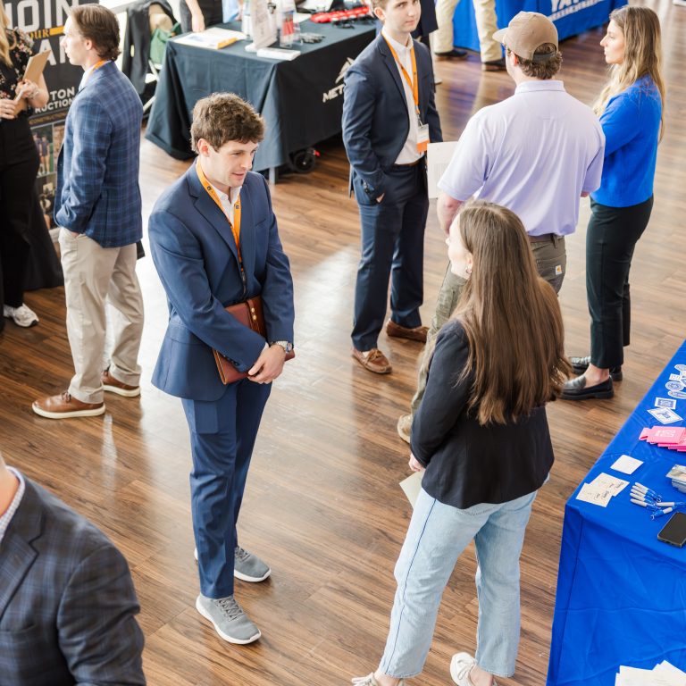 People in business attire converse and network at an indoor event with booths and tables displaying promotional materials.