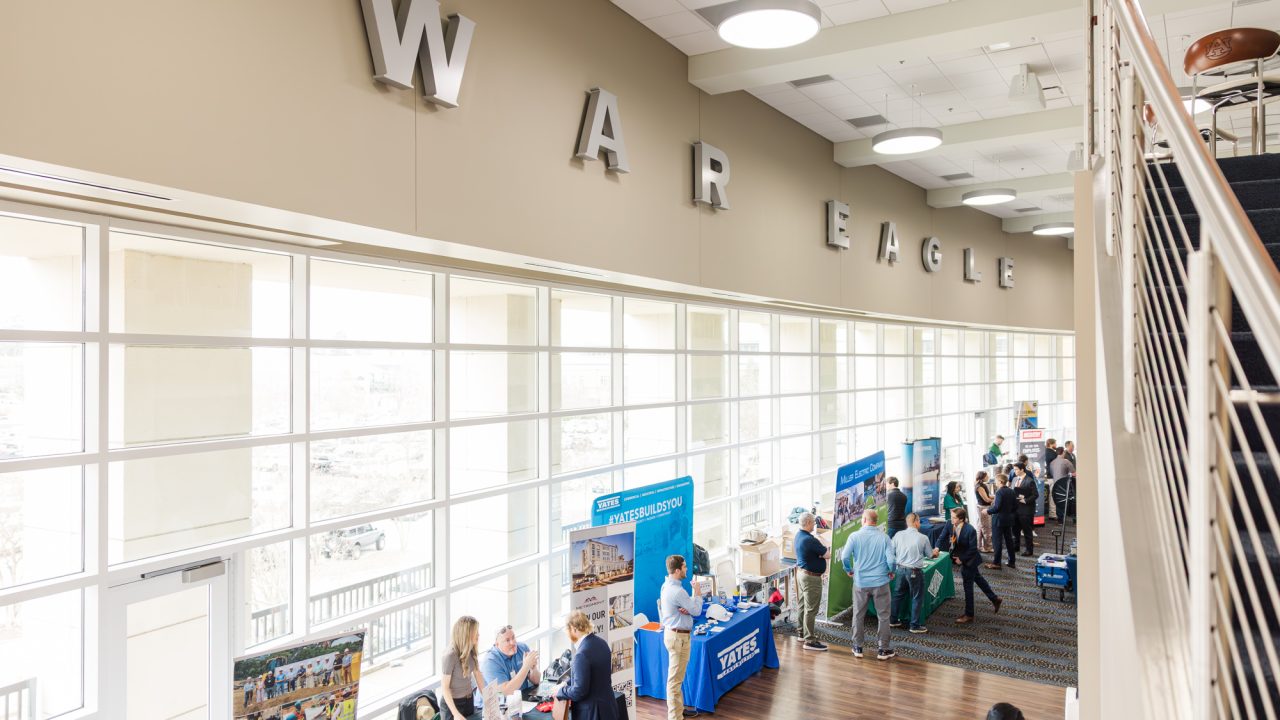 People visit booths at an indoor expo or career fair, with large WAR EAGLE letters mounted on the wall above the windows.