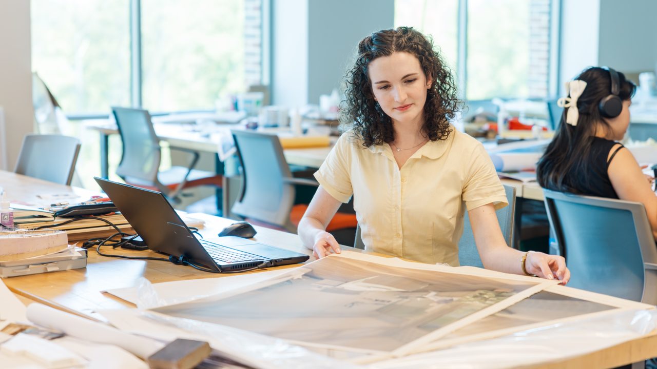 A woman in a yellow shirt examines a large printed image at a desk with a laptop; another person works at a separate desk in the background.