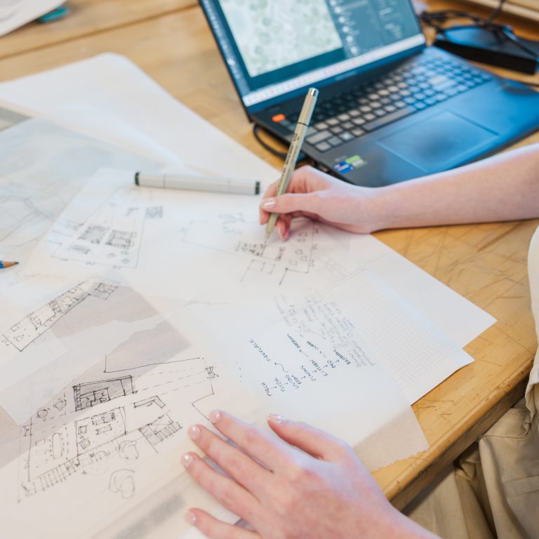Person working at a desk with architectural sketches, notes, and a laptop displaying a map or design software.