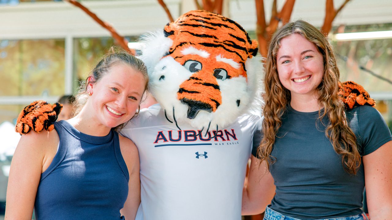 Two smiling people pose with the Auburn University tiger mascot outdoors, all standing close together in front of trees and a building.