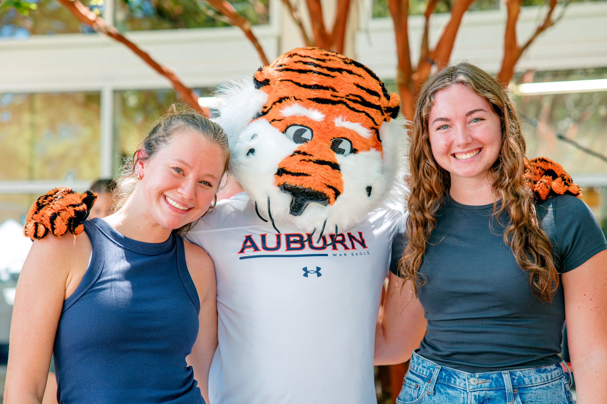 Two smiling people pose with the Auburn University tiger mascot outdoors, all standing close together in front of trees and a building.