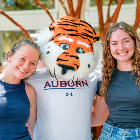 Two smiling people pose with the Auburn University tiger mascot outdoors, all standing close together in front of trees and a building.