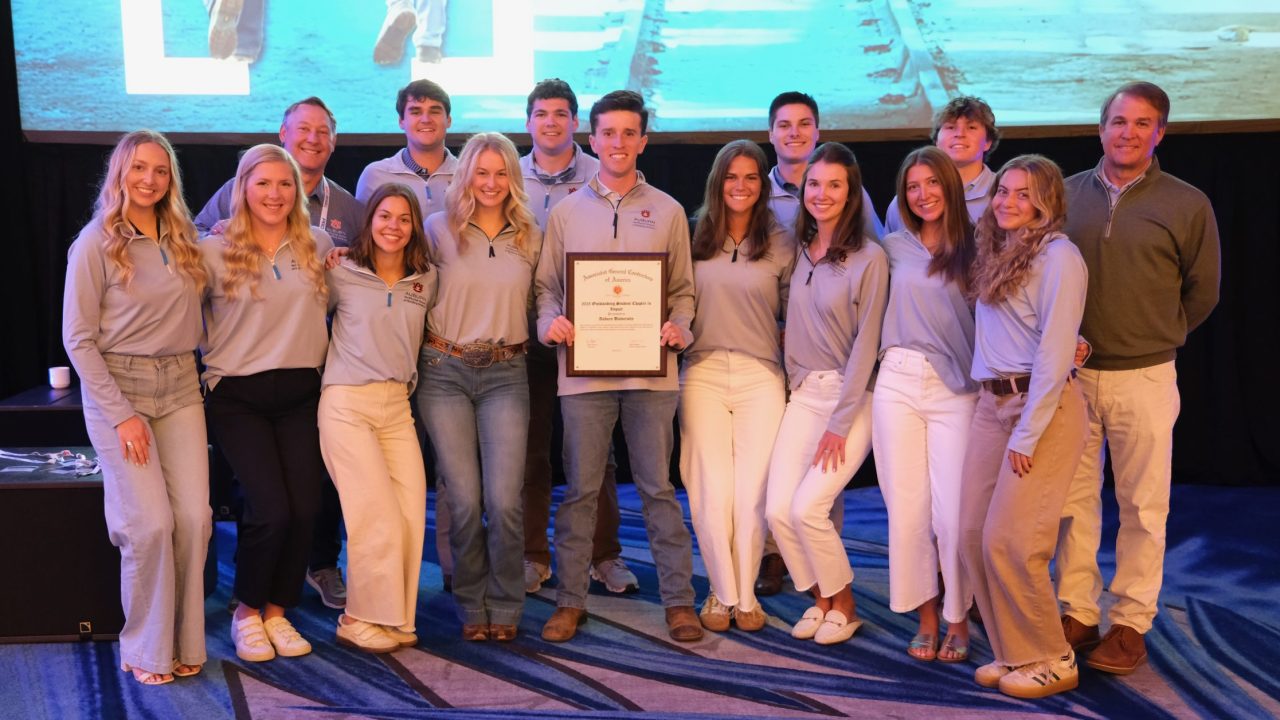 A group of fifteen young adults pose indoors, with one person in the center holding a framed certificate; a large screen is visible in the background.