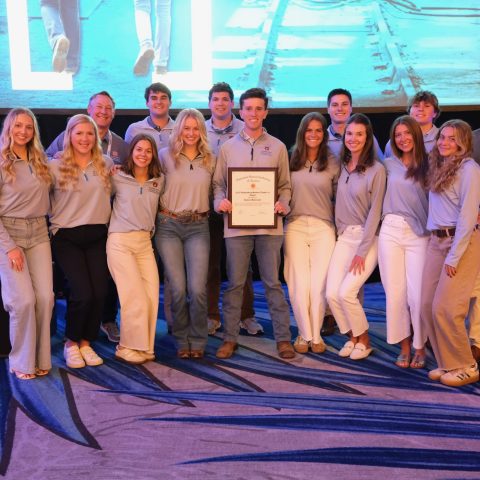 A group of fifteen young adults pose indoors, with one person in the center holding a framed certificate; a large screen is visible in the background.