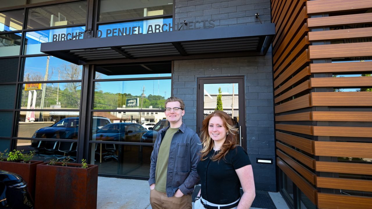 Two people stand outside the entrance of a building labeled BIRCHFIELD PENUEL ARCHITECTS with parked cars and trees visible in the background.