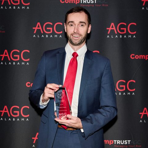A man in a blue suit and red tie holds an award in front of a backdrop with AGC Alabama and CompTrust AGC logos.