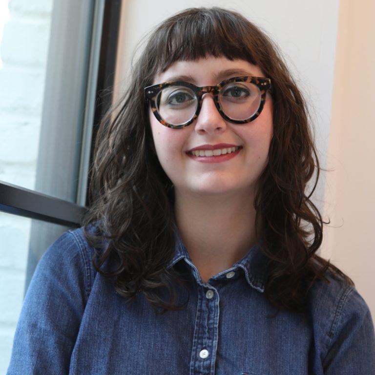 A woman with long brown hair and glasses, wearing a denim shirt, sits indoors near a window and smiles at the camera.