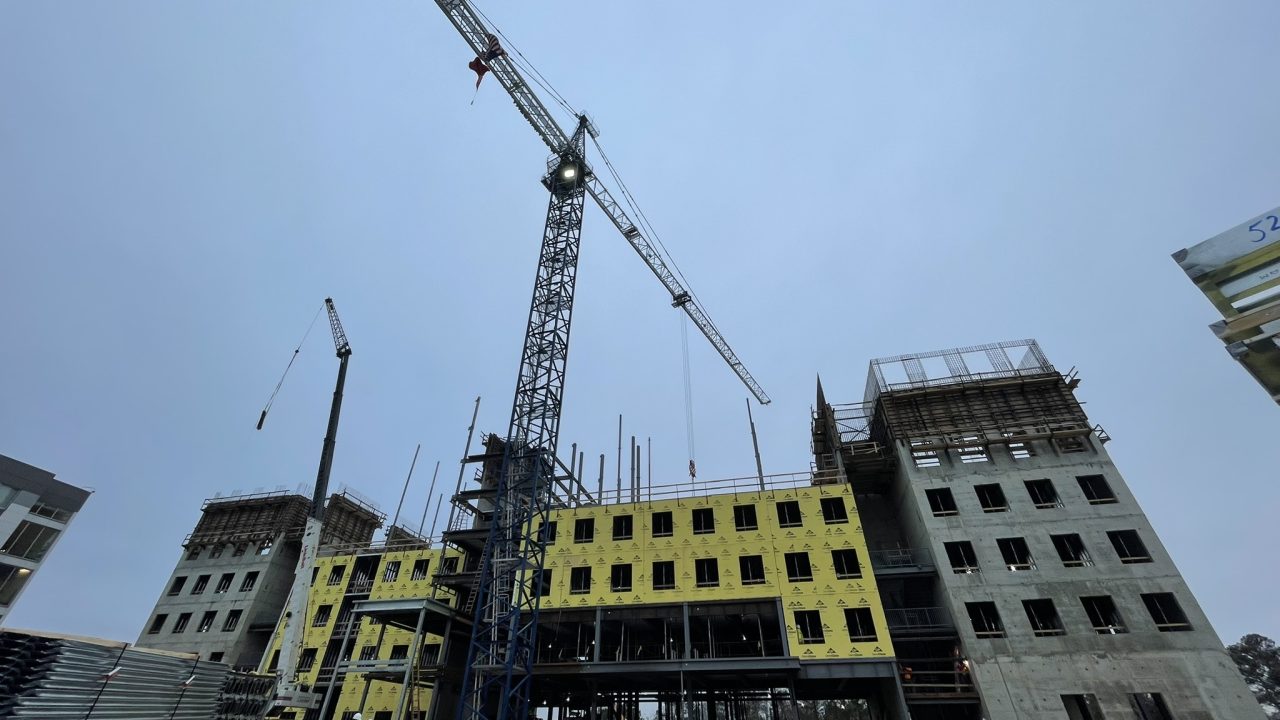 A multi-story building under construction with a large crane and exposed concrete; yellow insulation panels are visible on part of the structure.