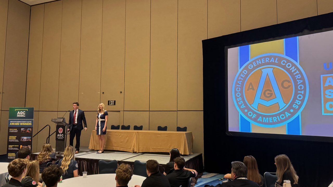 Two presenters stand at a podium on stage in front of an audience, with the Associated General Contractors of America logo displayed on a large screen.