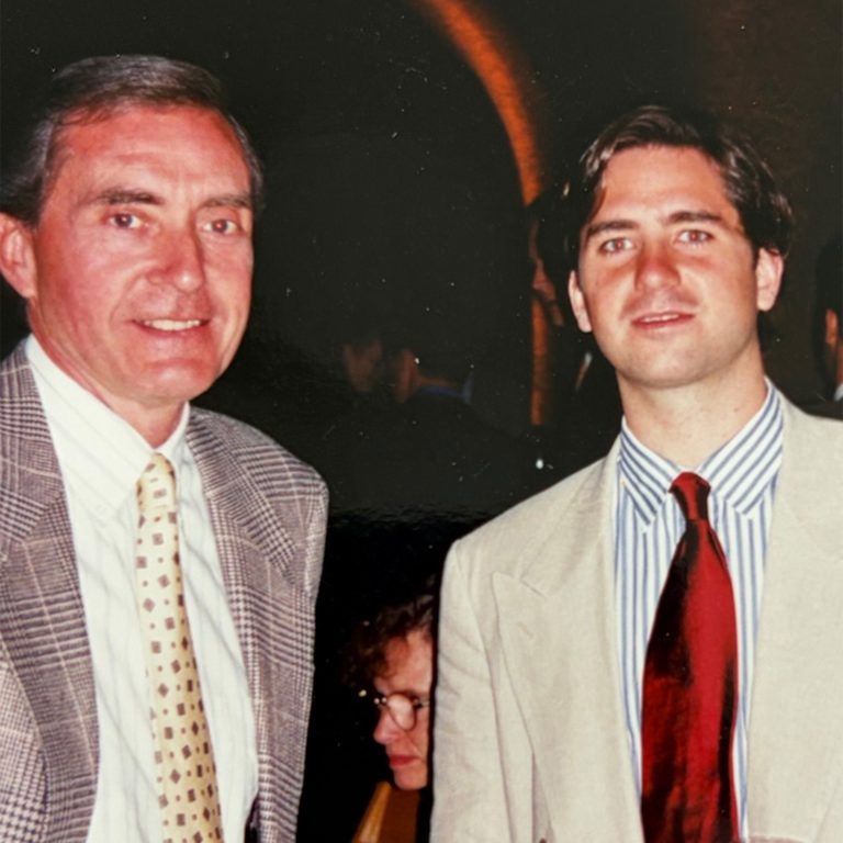 Two men wearing suits and ties pose together indoors at a formal event. A woman is seated in the background.