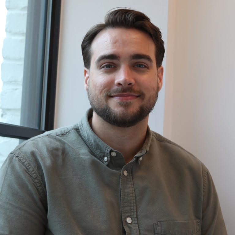 A man with short brown hair and a beard, wearing a green button-up shirt, sits indoors by a window and smiles at the camera.