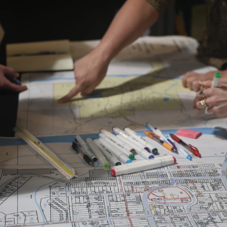 Two people review maps and documents on a table covered with markers, pens, rulers, and city planning blueprints.