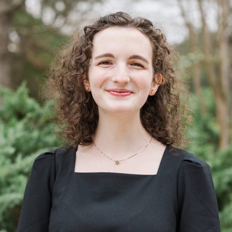 A woman with curly brown hair wearing a black dress stands outdoors in front of green trees, smiling at the camera.