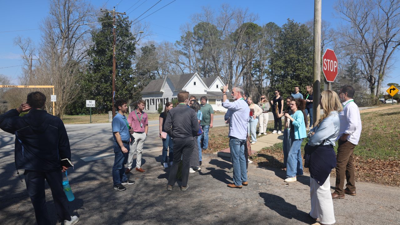 A group of people stand and talk at a street corner near a stop sign on a sunny day, with houses and trees in the background.