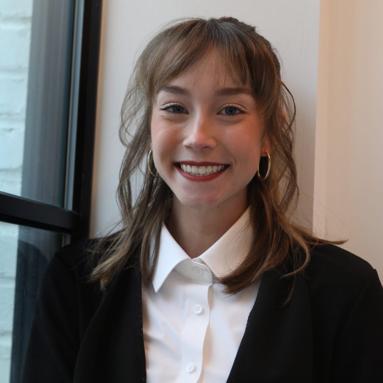 A woman with light brown hair and bangs, wearing a black blazer and white shirt, smiles while sitting by a window.