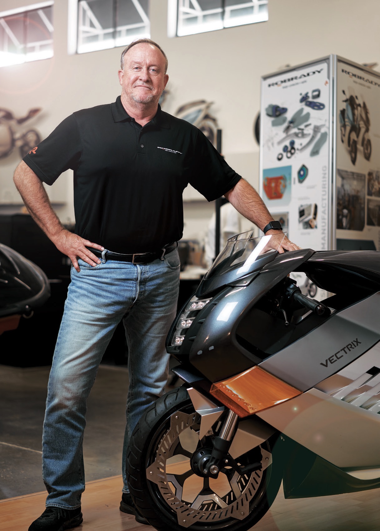 A man stands next to a Vectrix electric motorcycle inside a showroom or workshop, with posters and equipment visible in the background.