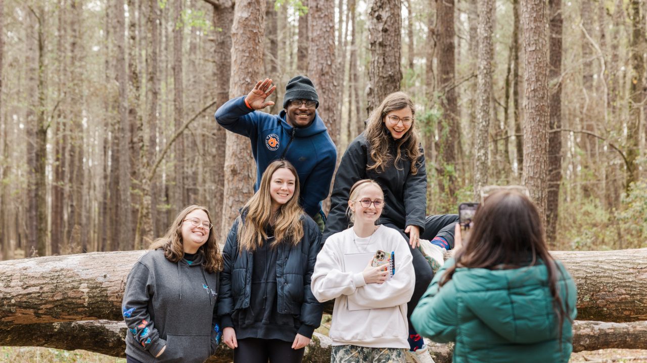 Five people pose for a photo on a fallen tree in a forest while a sixth person takes their picture; some are smiling and one waves at the camera.