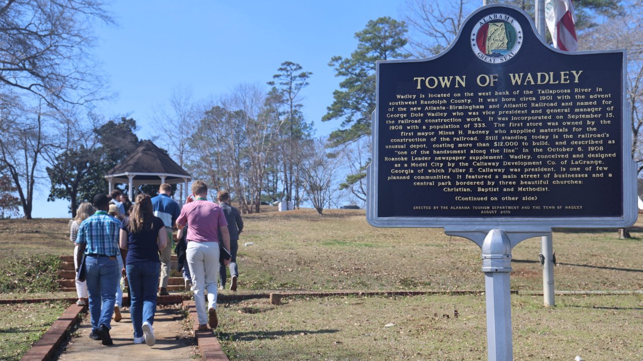 A group of people walk up a path toward a gazebo in a park near a historical marker sign for the Town of Wadley in Alabama.