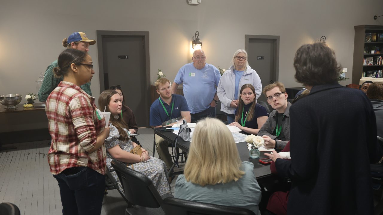 A group of people sit and stand around a table in a meeting room, engaged in discussion. Papers and drinks are on the table.
