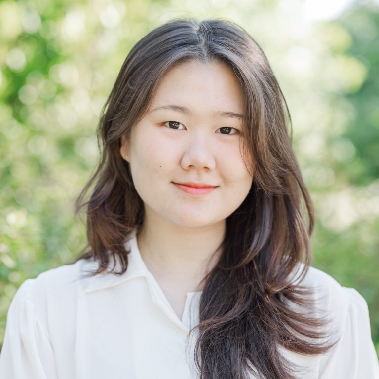 A young woman with long dark hair wearing a white blouse stands outdoors in front of greenery, looking at the camera with a neutral expression.