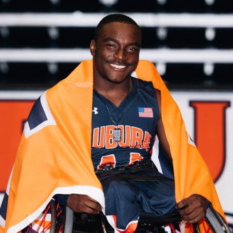 A wheelchair basketball player in an Auburn jersey smiles at the camera, draped in an orange and navy flag, with a basketball court backdrop.