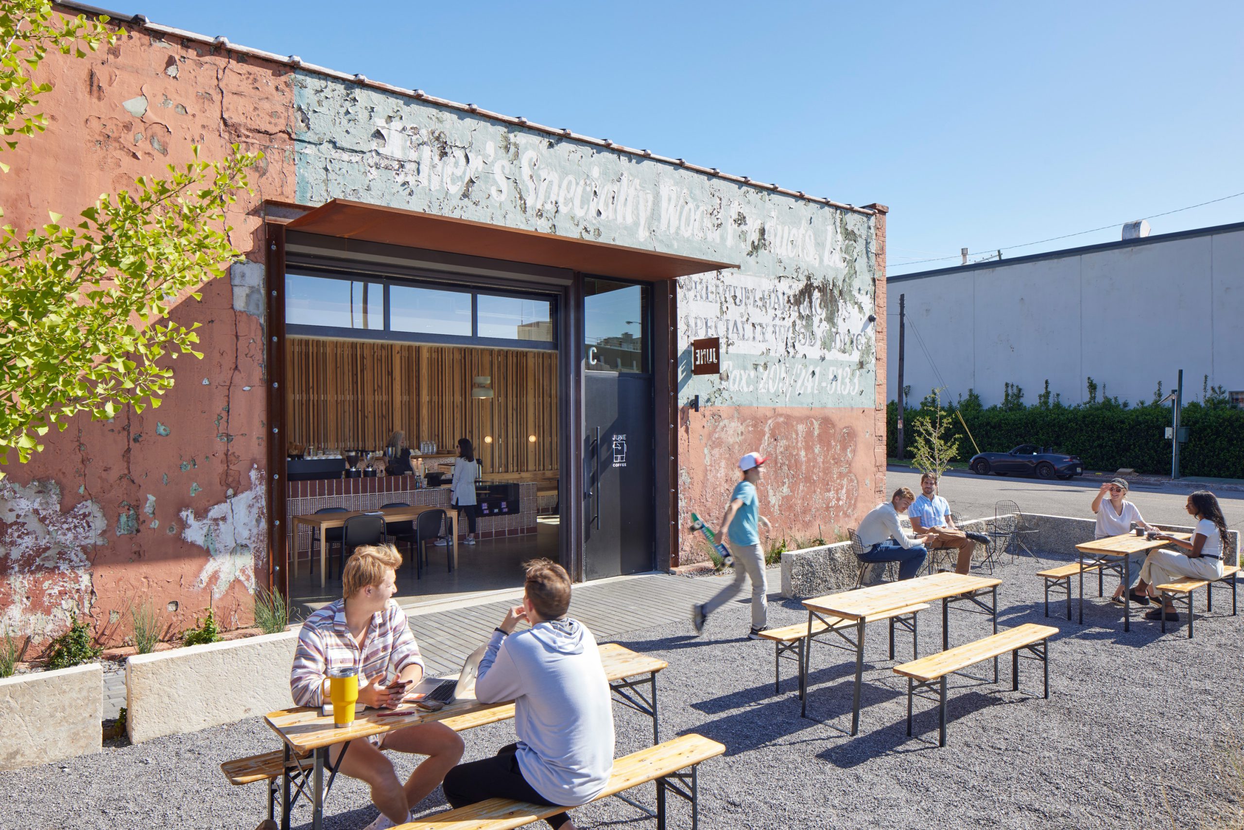 People sit at outdoor tables in front of a rustic, weathered café building on a sunny day; a few people walk nearby and the sky is clear.