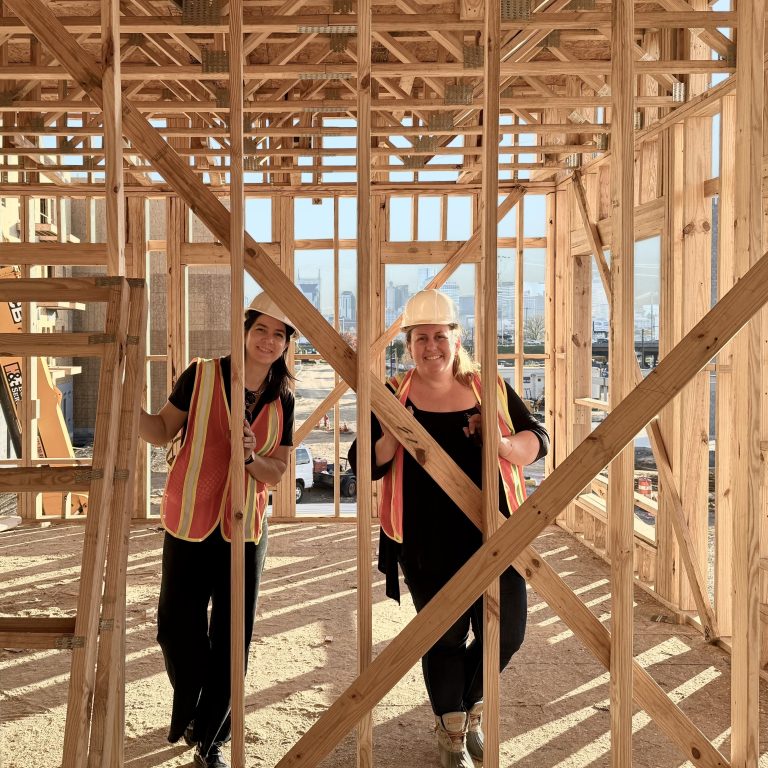 Two people wearing safety vests and hard hats stand inside the wooden frame of a building under construction, smiling at the camera.