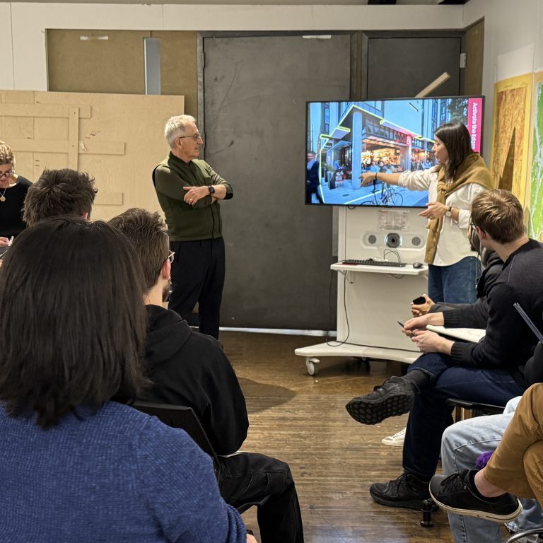 A woman points to a screen displaying a city street while presenting to a seated group in a classroom; a man stands nearby listening.
