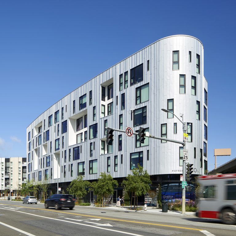Modern mid-rise apartment building on a city street corner with cars and a bus passing by under a clear blue sky.