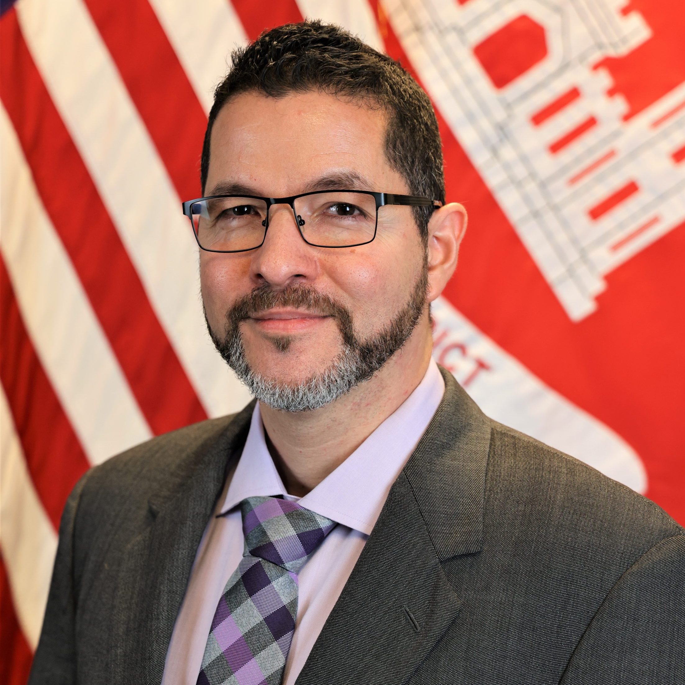 A man in a suit and glasses stands in front of U.S. and U.S. Army Corps of Engineers flags.