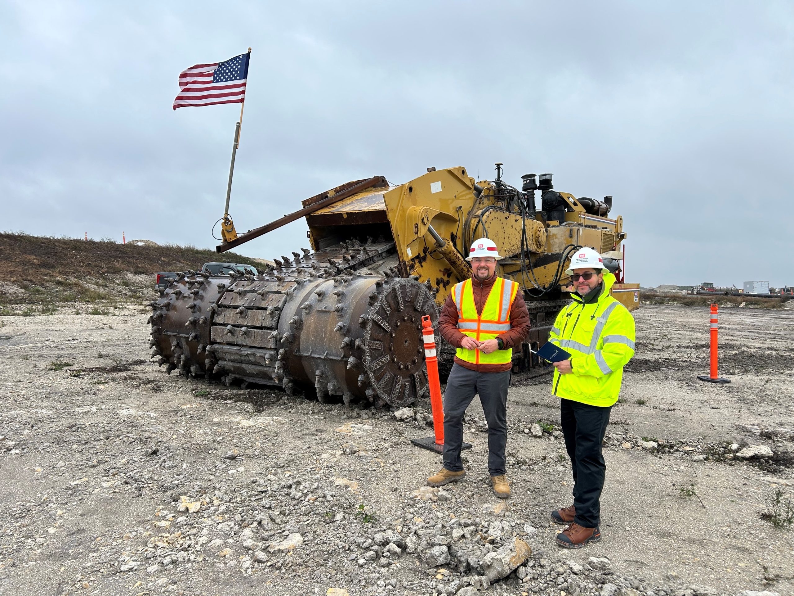 Two construction workers in safety gear stand in front of a large industrial machine with an American flag attached, on a gravel worksite under a cloudy sky.