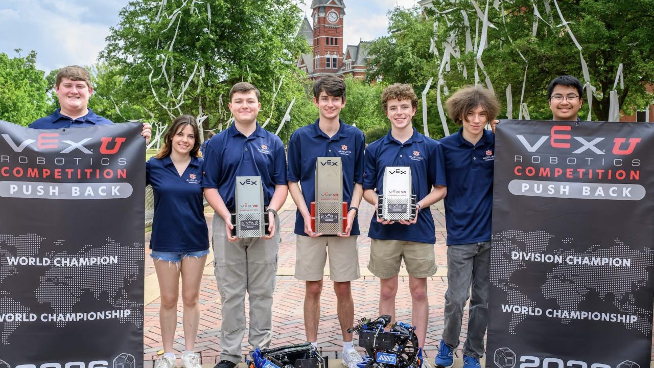 A group of seven students in matching shirts stand outdoors holding VEX Robotics trophies, flanked by two event banners, with robots on the ground in front of them.