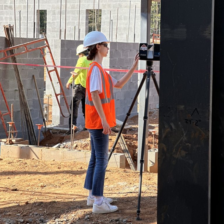 A woman in a hard hat and orange safety vest uses a surveying instrument on a construction site, with another worker and unfinished building in the background.
