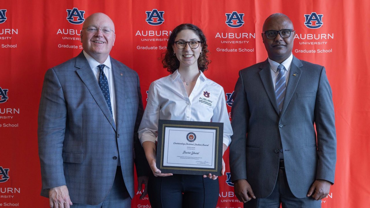Three people stand in front of an Auburn University Graduate School backdrop; the person in the center holds a framed certificate.
