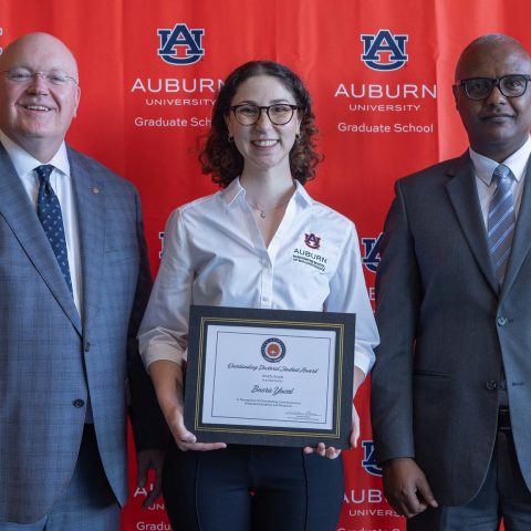 Three people stand in front of an Auburn University Graduate School backdrop; the person in the center holds a framed certificate.