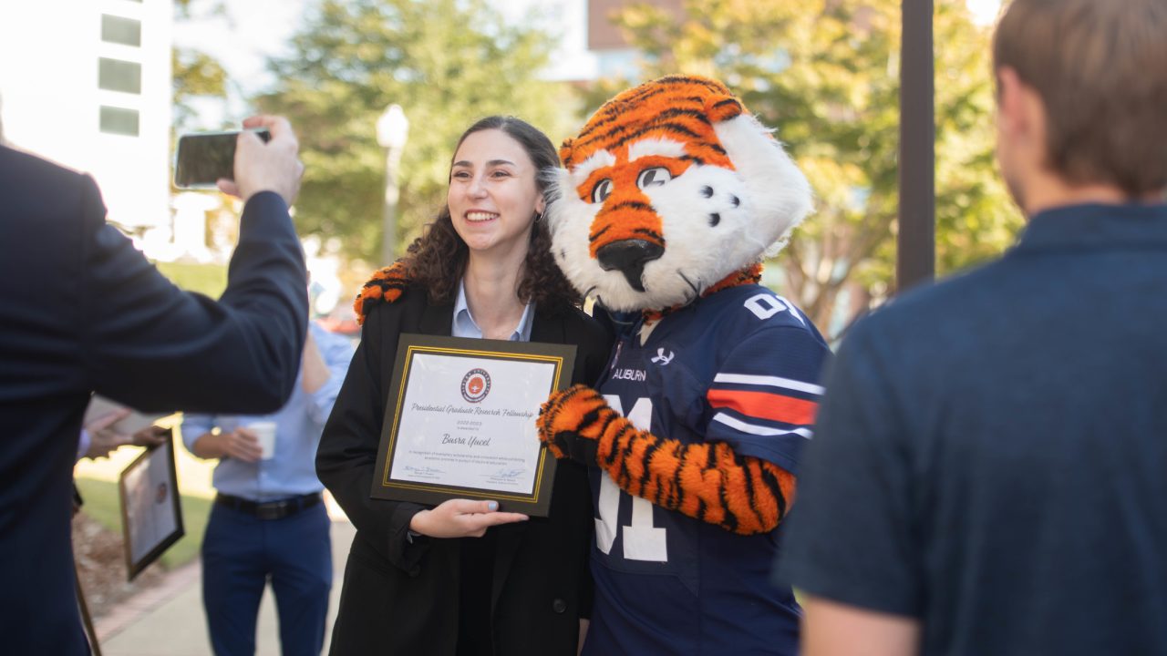 A woman holds a certificate and poses for a photo with a person in a tiger mascot costume outdoors, while others stand nearby.