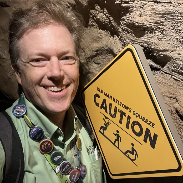 A man wearing a green shirt, lanyard, and buttons stands next to a yellow caution sign reading Old Man Keltons Squeeze with figures squeezing through a narrow passage.