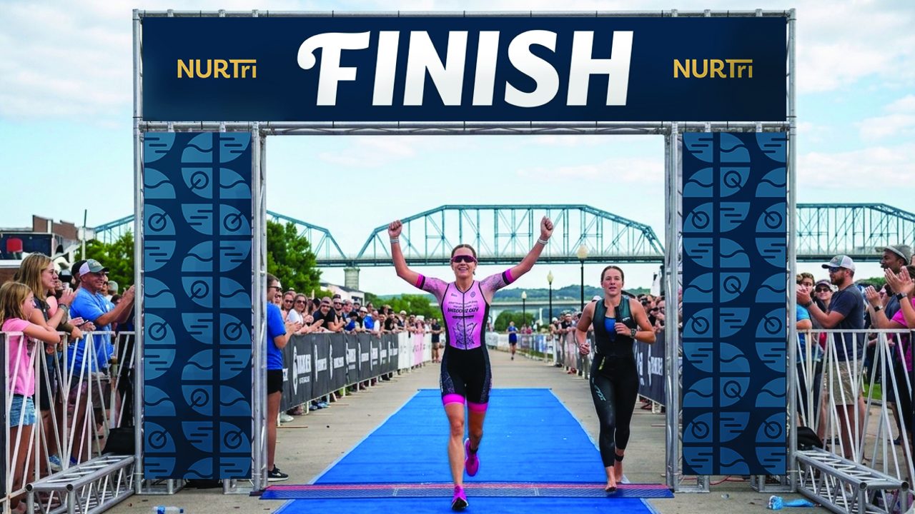 Two athletes cross the finish line at a triathlon event, with the leading athlete raising their arms in victory and spectators cheering from behind barriers.