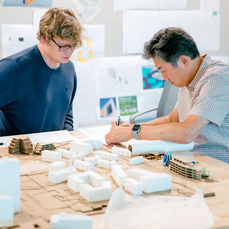 Two men sit at a table covered with architectural models and drawings, reviewing documents and making notes, with design plans posted on the wall behind them.