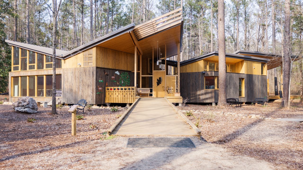 Modern wooden nature center with large angular roofs, surrounded by tall trees and forest, connected by a ramp and pathways.