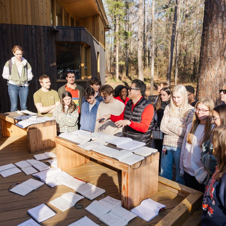 A group of people gathers outdoors around a table covered with open books and papers, listening to a person speaking. Trees and a wooden building are in the background.