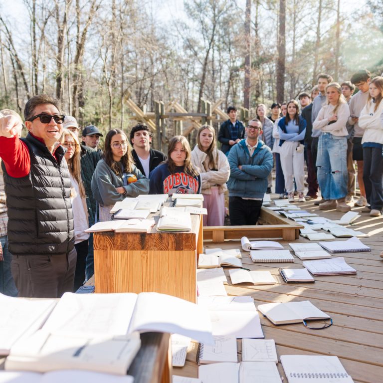 A person speaks to a large group gathered outdoors on a wooden deck, with notebooks and papers spread out on tables and the ground.