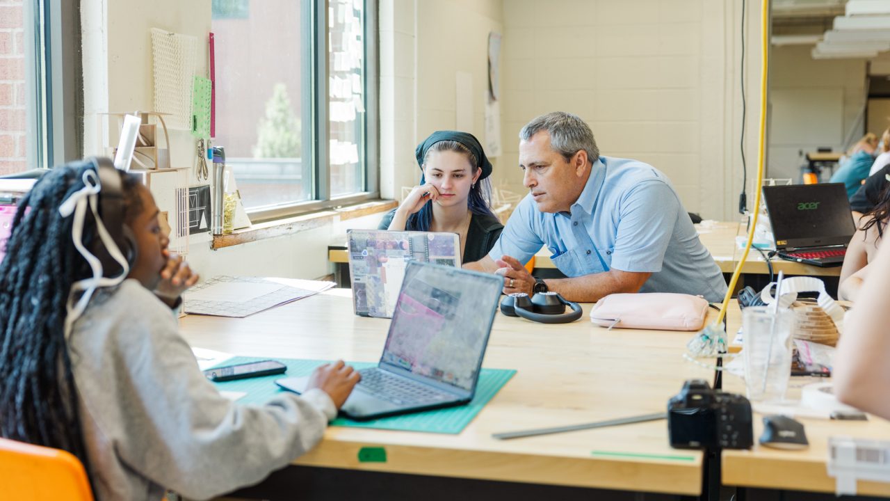 A teacher speaks with two students at a desk in a classroom with laptops and crafting materials on the table.
