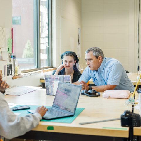 A teacher speaks with two students at a desk in a classroom with laptops and crafting materials on the table.