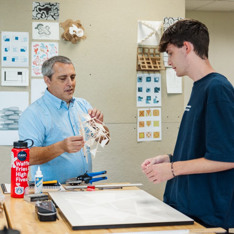 Two people in a classroom work on a wooden model at a table, with art and design sketches displayed on the wall behind them.