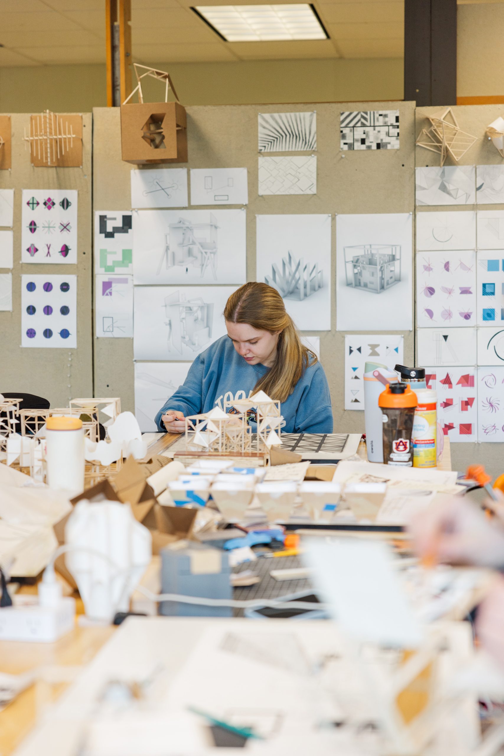 A person works on architectural models at a cluttered desk in a studio, with sketches, diagrams, and models displayed on the wall behind.